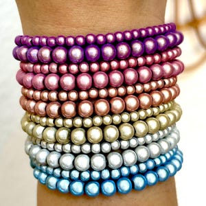 Könnte beinhalten: Ein Stapel aus zehn Perlenarmbändern mit einem Farbverlauf. Die Armbänder reichen von Dunkelviolett oben über Rosa-, Kupfer-, Gold- und Silbertöne bis hin zu hellem Blau unten. Jedes Armband besteht aus runden Perlen in einer Reihe.