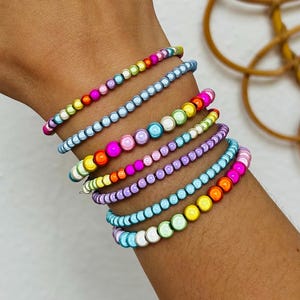 Könnte beinhalten: Ein Stapel aus sieben Perlenarmbändern mit kleinen, runden, glänzenden Perlen in einer Vielzahl leuchtender Farben, darunter Pink, Blau, Gelb, Grün, Orange und Violett. Die Armbänder sind in einem geschichteten Muster am Handgelenk angeordnet.