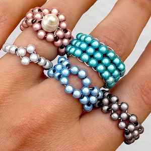Könnte beinhalten: Eine Sammlung von vier Perlenringen, die an den Fingern getragen werden. Die Ringe bestehen aus kleinen, runden Perlen mit Metallic-Finish in Pink, Türkis, Hellblau und Dunkelgrau. Ein pinkfarbener Ring ist mit einer größeren weißen Perle verziert.