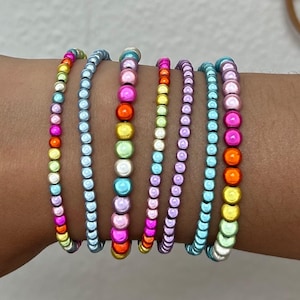 Könnte beinhalten: Eine Sammlung von Armbändern mit Perlen in verschiedenen Farben, darunter Rosa, Orange, Gelb und Blau. Die Armbänder bestehen aus kleinen, runden Perlen, die zu einem farbenfrohen und stilvollen Accessoire aufgereiht sind.