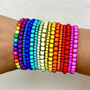 Könnte beinhalten: Ein Stapel aus mehreren elastischen Armbändern, bestehend aus kleinen, runden, glänzenden Perlen in einem lebendigen Regenbogen-Farbverlauf, der von Violett und Blau über Grün, Gelb, Orange und Rot bis hin zu Pink reicht.