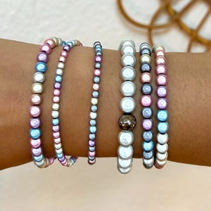 Könnte beinhalten: Eine Sammlung von sechs Armbändern mit Perlen in verschiedenen Stilen und Farben. Die Armbänder haben schillernde Perlen in Rosa-, Blau- und Silbertönen. Ein Armband hat eine einzelne braune Perle.