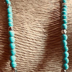 May include: A long necklace featuring round, turquoise-colored beads with dark veining, interspersed with small, metallic, faceted silver-toned cube beads. The beads are arranged in a single strand, creating a uniform pattern throughout the length.