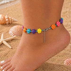 May include: A vibrant anklet with flower-shaped beads in orange, turquoise, purple, and yellow. The anklet is displayed on an ankle on a sandy beach, with seashells and a starfish visible in the background.