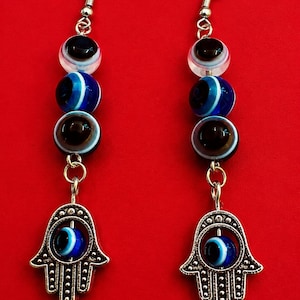 May include: A pair of dangle earrings featuring silver-toned metal hooks and three stacked glass beads in shades of black, white, and blue. Each earring terminates in a silver-toned metal hamsa charm containing a small blue and white evil eye bead.