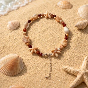 Peut inclure: Un bracelet sur le thème de la plage avec des coquillages, des étoiles de mer et de petites perles rouges. Le bracelet est composé de différentes formes et tailles de coquillages, avec un fermoir argenté et une chaîne d'extension. Il est posé sur du sable.