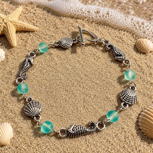 May include: A silver-toned chain bracelet featuring alternating charms of seahorses and seashells, interspersed with translucent light blue beads. The bracelet is secured with a heart-shaped toggle clasp, creating a cohesive nautical-themed accessory.