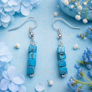 May include: A pair of turquoise cube earrings with silver-colored hooks. Each earring features five turquoise cubes stacked vertically. The earrings are displayed on a light blue surface with blue flowers and small white flowers.