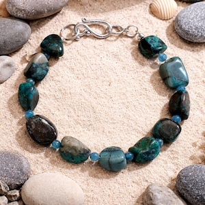 May include: A beaded bracelet featuring large, irregularly shaped turquoise and dark green stones. Small blue beads separate the larger stones. The bracelet is secured with a silver-colored clasp. The jewelry rests on a sandy surface.