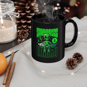 May include: Black ceramic mug with a green alien design and the text "SUCCESS IS OUT OF THIS WORLD" and "UNITED". The alien is on a spaceship. The mug has a curved handle. The image includes festive decorations.