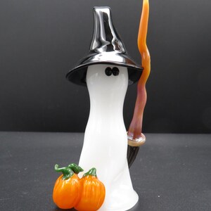 Peut inclure: Une figurine en verre représentant un fantôme blanc portant un chapeau pointu noir, tenant un grand bâton ondulé orange et marron. Deux petites citrouilles orange à tiges vertes sont placées à la base du fantôme aux deux yeux noirs.