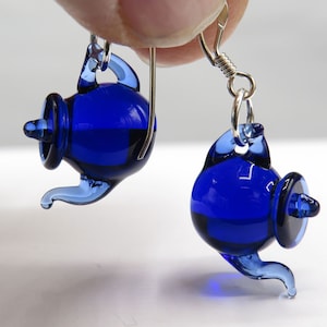Op de afbeelding: Een paar oorbellen in de vorm van theepotten van kobaltblauw glas. Elke oorbel heeft een miniatuur, doorschijnende blauwe glazen theepotbedel met een gebogen tuit en een klein deksel, hangend aan een zilverkleurige metalen oorhaak.