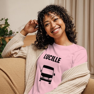 Può includere: Una maglietta rosa chiaro a maniche lunghe con il nome "LUCILLE" in lettere nere in grassetto e una grafica di autobus nera. La maglietta è realizzata in un materiale morbido.