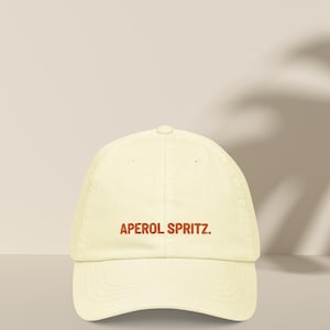 Könnte beinhalten: Eine hellgelbe Baseballkappe mit dem Schriftzug "APEROL SPRITZ." in Orange gestickt.