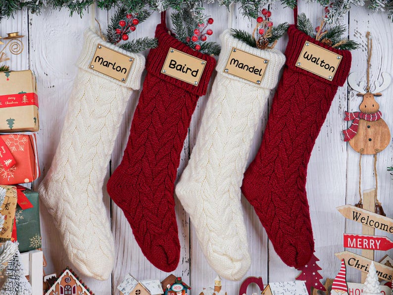 May include: Four personalized Christmas stockings in cream and red cable knit, each with a wooden name tag. The names are Marlon, Baird, Mandel, and Walton. Festive holiday decor with greenery and a wooden snowman.