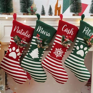 May include: Four personalized Christmas stockings with red and green stripes. The stockings are decorated with a snowflake, paw print, and a Christmas tree. The names Natalie, Mona, Cash, and Pete are written on the stockings.