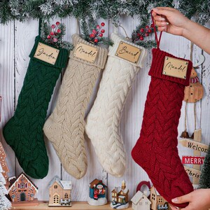 May include: Four cable-knit Christmas stockings in green, beige, white, and red, each with a wooden name tag. The red stocking is held up by a hand. Festive holiday decor includes miniature houses and a Christmas tree.