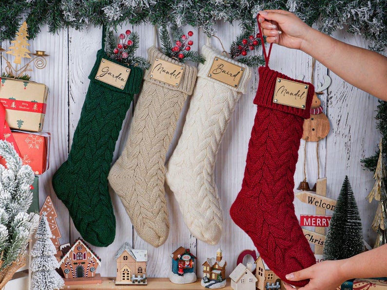 May include: Four cable-knit Christmas stockings in green, beige, white, and red, each with a wooden name tag. The red stocking is held up by a hand. Festive holiday decor includes miniature houses and a Christmas tree.
