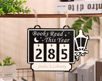 May include: Black and white wooden sign with the text "Books Read This Year" and the number "285". The sign includes a decorative lantern and the name "Jodie's Library". It hangs by a natural twine.