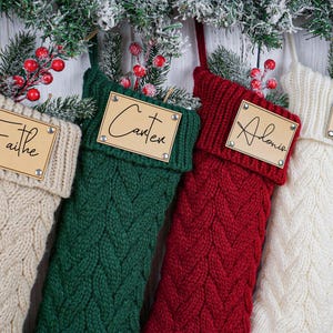 以下が含まれることがあります： クリーム、緑、赤、白のケーブルニットのクリスマスストッキング4つ。それぞれに「Faithe」、「Carter」、「Alonie」、「Sandra」と名前が書かれた木製のネームタグが付いています。お祝いのホリデーデコレーション。