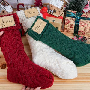 May include: Three cable-knit Christmas stockings in red, white, and green. Each stocking has a tag with a name on it. The red stocking is held by a hand. The stockings are on a wooden surface with Christmas decorations.