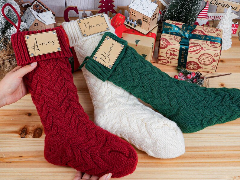 May include: Three cable-knit Christmas stockings in red, white, and green. Each stocking has a tag with a name on it. The red stocking is held by a hand. The stockings are on a wooden surface with Christmas decorations.
