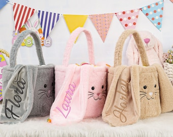 Könnte beinhalten: Drei flauschige Osterhasen-Körbe in Grau, Rosa und Beige. Jeder Korb hat einen Griff und ein Hasengesicht mit gestickten Schnurrhaaren. Die Namen "Flora", "Laura" und "Harley" sind auf den Ohrenklappen gestickt. Bunte Osterdekorationen im Hintergrund.