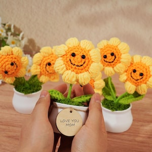 May include: Crocheted yellow sunflowers with smiling faces in white pots. The flowers have orange centers and green leaves. A wooden tag reads "LOVE YOU MOM".