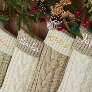 May include: Four cream and beige cable-knit Christmas stockings, each embroidered with a name in gold or white. The background is decorated with festive greenery and ornaments.