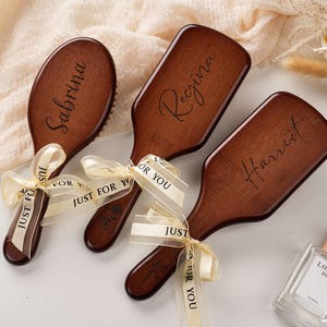 Könnte beinhalten: Drei personalisierte Holzbürsten mit den Namen "Sabrina", "Regina" und "Harriet". Jede Bürste ist mit einem cremefarbenen Band mit der Aufschrift "Just For You" versehen. Eine Parfümflasche ist ebenfalls im Bild.
