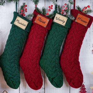 May include: Four personalized Christmas stockings in green and red, with names on leather tags. The stockings are knit with a cable pattern and decorated with sprigs of berries. A wooden reindeer ornament hangs nearby.