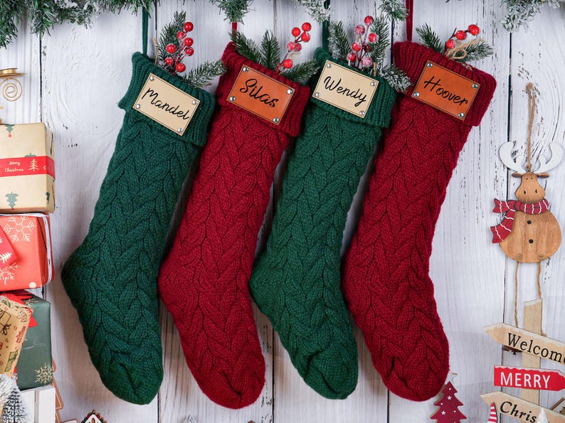 May include: Four personalized Christmas stockings in green and red, with names on leather tags. The stockings are knit with a cable pattern and decorated with sprigs of berries. A wooden reindeer ornament hangs nearby.