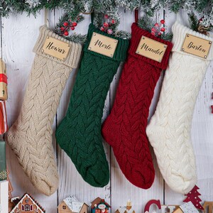 May include: Four cable-knit Christmas stockings in beige, green, red, and white hang against a white wood background. Each stocking has a wooden name tag. Festive decorations include a wooden reindeer ornament and wrapped gifts.
