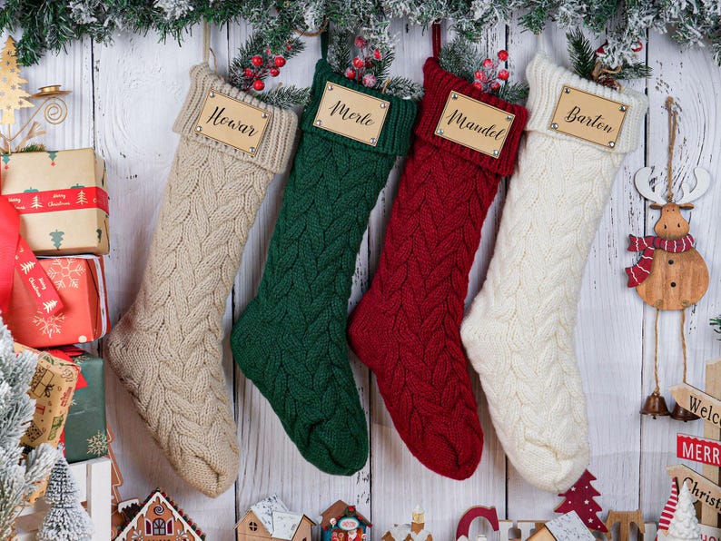 May include: Four cable-knit Christmas stockings in beige, green, red, and white hang against a white wood background. Each stocking has a wooden name tag. Festive decorations include a wooden reindeer ornament and wrapped gifts.