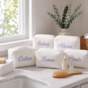 May include: Five white waffle-textured fabric pouches with blue embroidered names including Sidney, Alexia, Cotton, Leona, and Paula. The rectangular bags feature a zipper closure and are arranged together on a white marble surface.