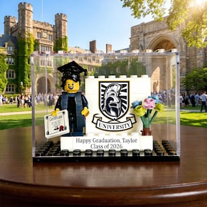 May include: A Lego graduation display featuring a minifigure in a cap and gown holding a certificate. The display includes a university crest, flowers, and the text "Happy Graduation, Taylor Class of 2026" inside a clear case.