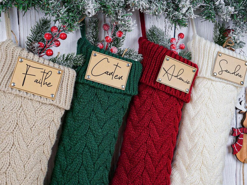 May include: Four cable-knit Christmas stockings in cream, green, red, and white, each with a wooden name tag. The names "Faithe", "Carter", "Alonie", and "Sandra" are written on the tags. Festive holiday decor.