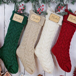 May include: Four cable-knit Christmas stockings in green, beige, white, and red, each with a wooden tag that reads "Mandel." The stockings are hanging against a white wooden background, decorated with greenery and berries.