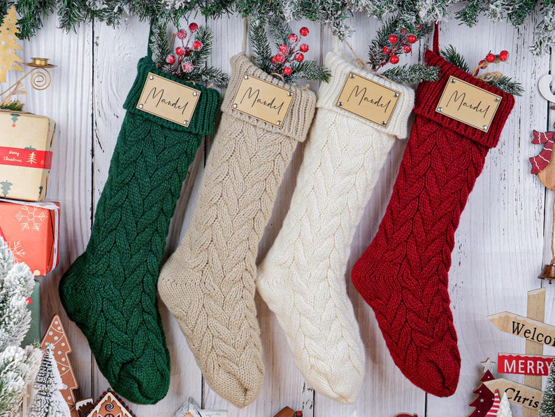 May include: Four cable-knit Christmas stockings in green, beige, white, and red, each with a wooden tag that reads "Mandel." The stockings are hanging against a white wooden background, decorated with greenery and berries.