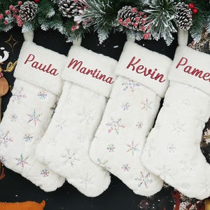 May include: Four white Christmas stockings with names embroidered in red: Paula, Martina, Kevin, and Pamela. The stockings have a snowflake pattern and are hanging from a garland of pine and berries.