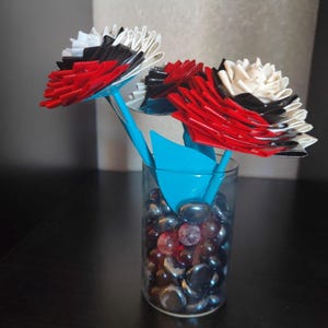 May include: Three red, white, and black duct tape flowers in a clear glass vase with blue glass stones.