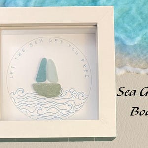 Könnte beinhalten: Ein weiß gerahmtes Kunstwerk mit einem Schiff aus Meerglas und dem Text "Lass das Meer dich frei sein". Das Schiff besteht aus drei Teilen Meerglas, zwei blauen und einem grünen. Der Hintergrund ist weiß mit einem blauen Wellendesign.