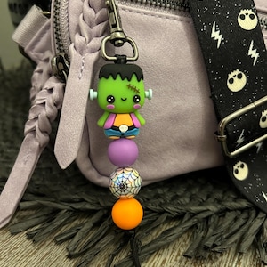 May include: A Halloween-themed keychain charm featuring a green Frankenstein's monster figure, purple, white, and orange beads, and a black tassel. The charm is attached to a lavender purse with a black and white skull strap.