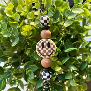 May include: A black and white checkered bead charm with a brown smiley face bead and brown wooden beads. The charm is attached to a black metal clip.