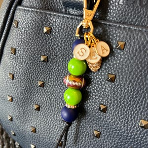 May include: A gold-toned keychain with a navy blue, green, and football-themed bead design. The keychain includes charms with the letters "SEA" and a small, jeweled charm. The keychain is attached to a navy blue handbag with gold studs.