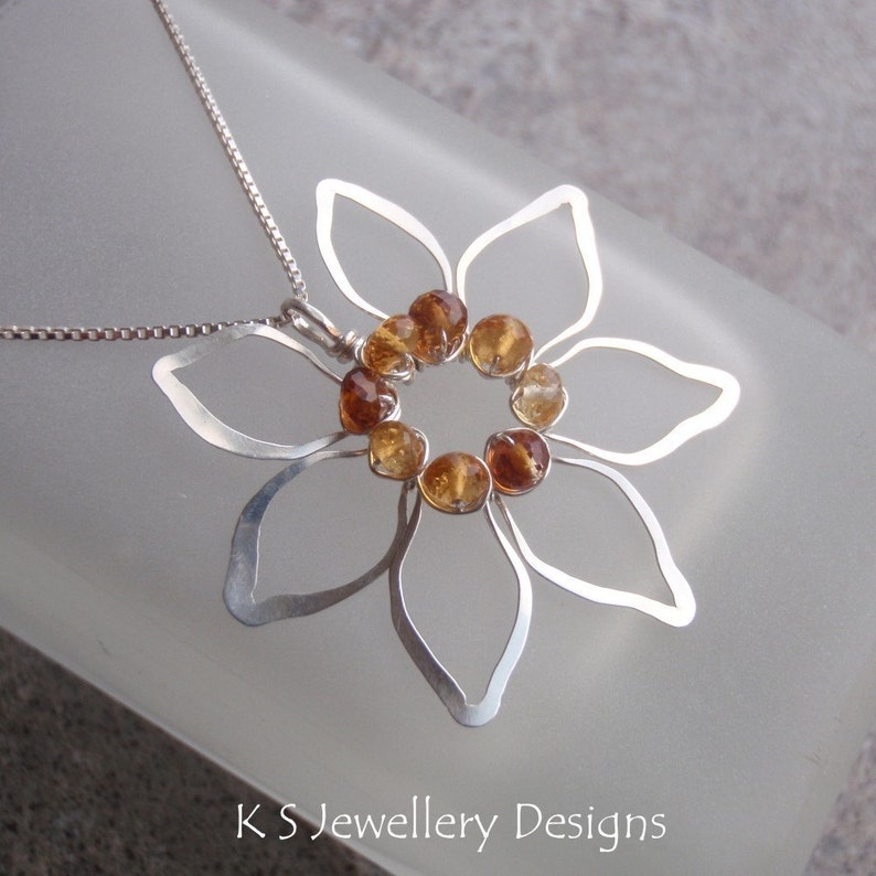 May include: A silver-toned pendant necklace featuring a stylized flower design. The flower has openwork petals and a center cluster of amber-colored gemstones. The necklace is on a silver chain. The text "KS Jewellery Designs" is at the bottom.