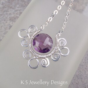 May include: A silver necklace with a purple amethyst gemstone pendant. The pendant is surrounded by silver wire that is twisted into a decorative design.