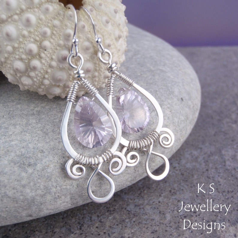 May include: Silver dangle earrings with pink teardrop-shaped crystals and silver wire detailing. The earrings are on a gray stone surface.