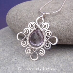 May include: A silver wire wrapped pendant featuring a light purple amethyst gemstone. The pendant is in the shape of a stylized flower with intricate swirls.