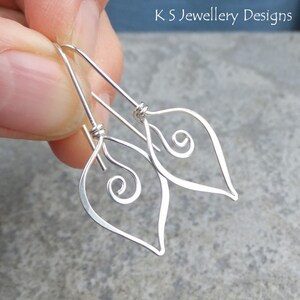 May include: A pair of silver leaf-shaped earrings. Each earring features a delicate wire outline with a curled spiral design inside the leaf shape. The earrings are held against a gray background. The text "K S Jewellery Designs" is visible.